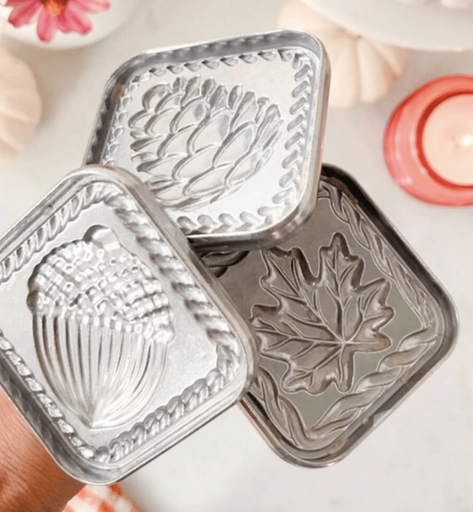 Three metallic cookie cutters with embossed designs of a pinecone, an acorn, and a maple leaf are held in hand. A pink flower and a lit candle blur in the background, offering a cozy atmosphere perfect for crafting Halloween treats for kids.